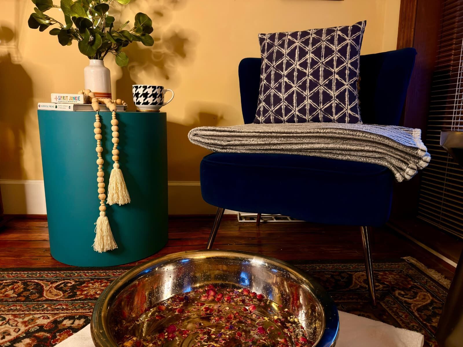 A warm foot bath scattered with rose petals before a blue velvet armchair, a wool throw over its arm.
