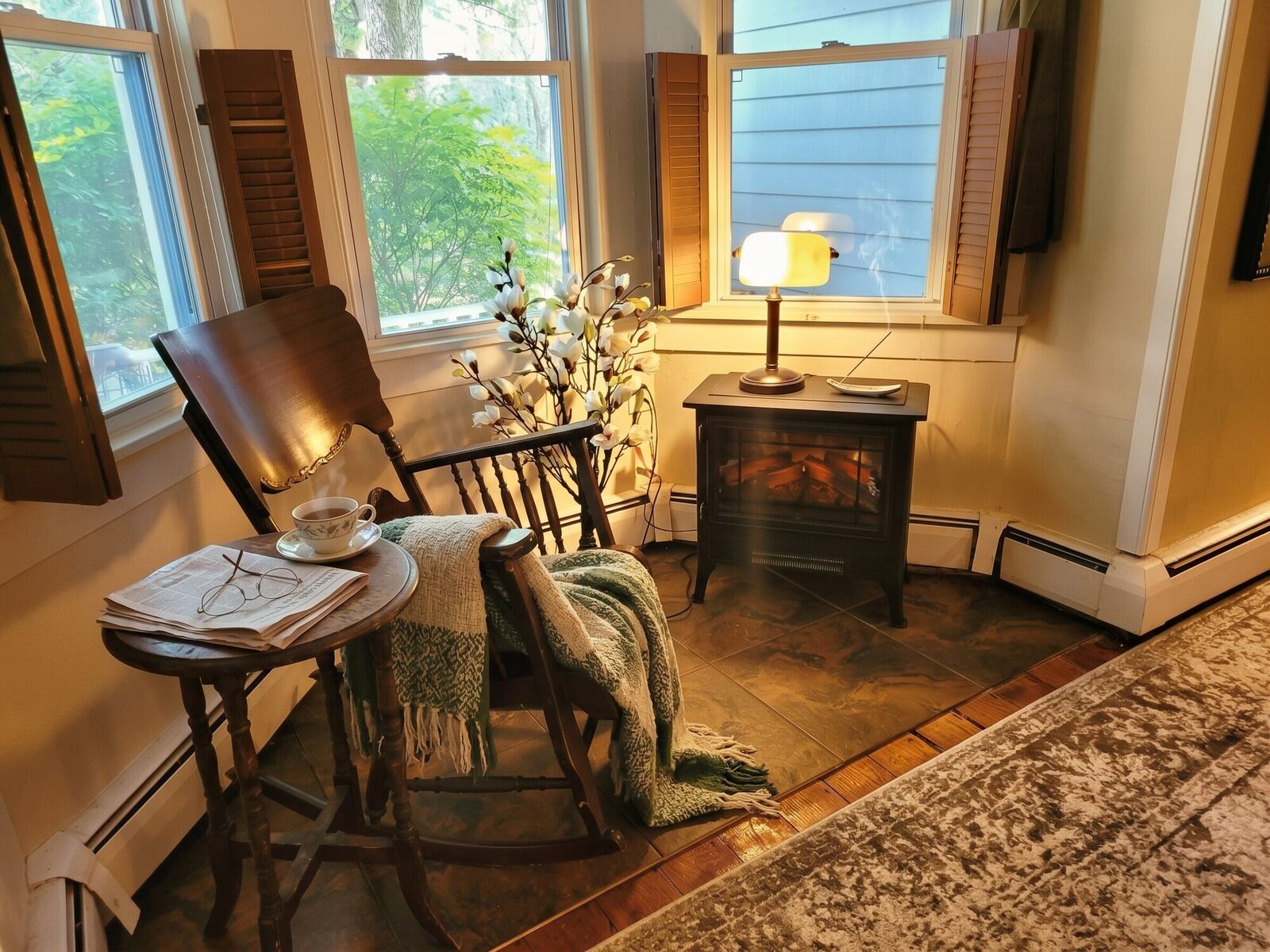 A rocking chair by the window in the Pump House front foyer, a blanket over its arm, a lamp lit beside it and a small stove in the corner.
